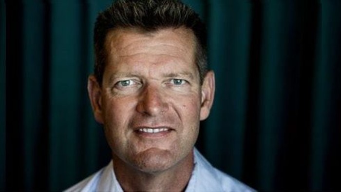 En mand med kort hår og briller, smilende foran mørkt tæppe.