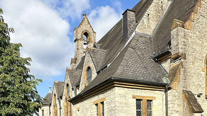 Historische Steinkirche mit Spitzbögen, steilem Dach und angrenzenden Bäumen unter einem teilweise bewölkten Himmel.