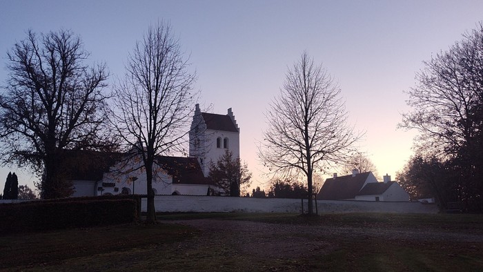 En lille kirke med tårn og et par andre bygninger i en åben landskab med træer og en blå himmel.