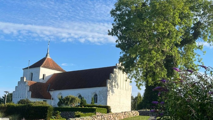 En hvid kirke med rødt tag og tårn ved en grøn plads med træer og blomster.