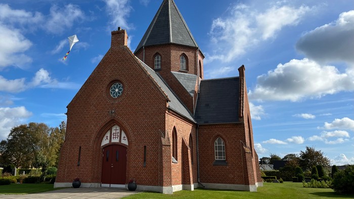 En rød murstenskirke med spids tårn og klokke under et blødt blå himmel.
