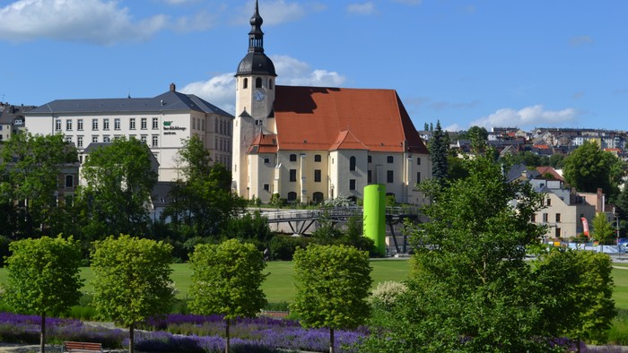 Ein großer weißer Gebäudekomplex mit rotem Dach und Turm, umgeben von grünen Bäumen und blühenden Pflanzen, unter einem klaren blauen Himmel