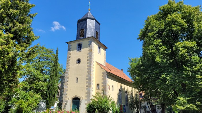 Kirchengebäude mit Turm und Uhr, umgeben von Bäumen unter blauem Himmel