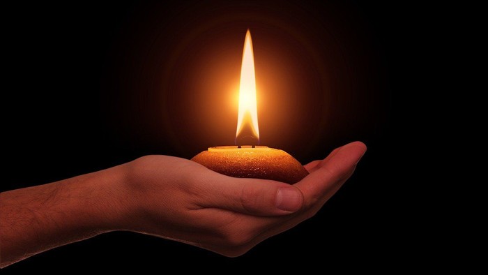 Hand hält brennende Kerze mit sichtbarer Flamme vor schwarzem Hintergrund.