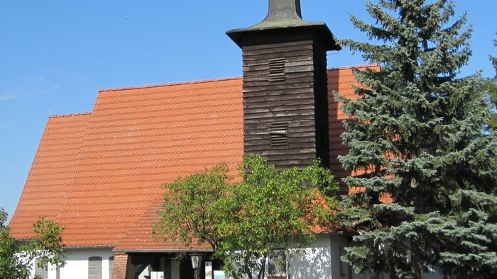 Kirchengebäude mit hohem Turm und rotem Dach, umgeben von Bäumen und Büschen