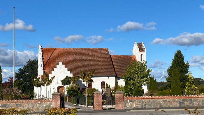 En hvid kirke med rødt tag og murstenet indhegning under et blødt blå himmel.