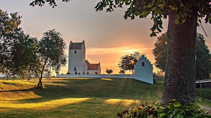 En lille kirke med tårn og et sidebygning på en grøn eng med træer og solnedgang