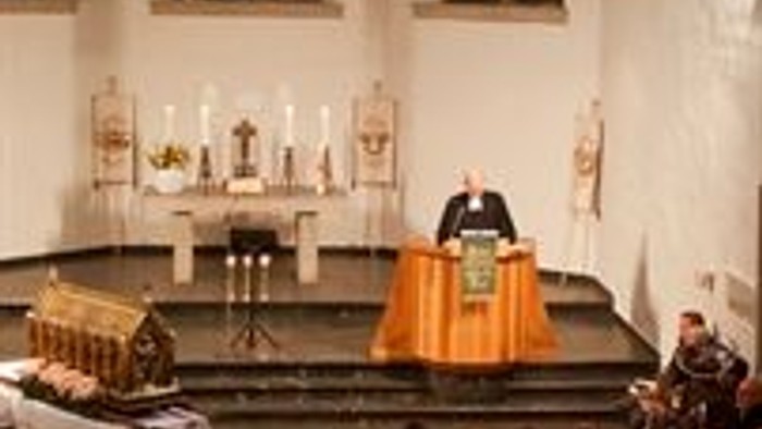 Ein feierlicher Gottesdienst zeigt einen Redner am Pult mit einem Publikum, das in Kirchenbänken sitzt.