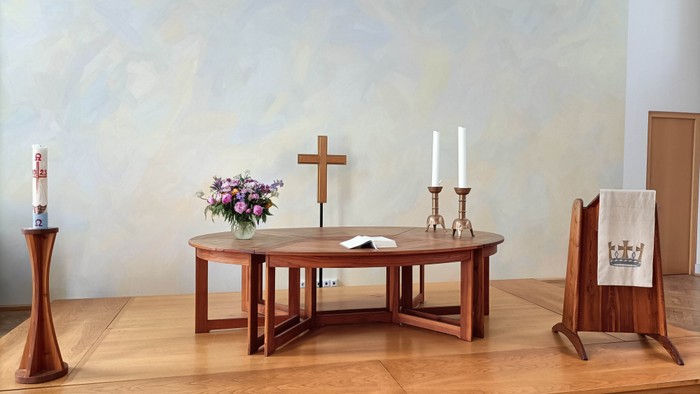 Ein ruhiger Kapellenaltar mit Holztisch, Kerzen, Kreuz, Blumen und einem brennenden Kerzenständer.