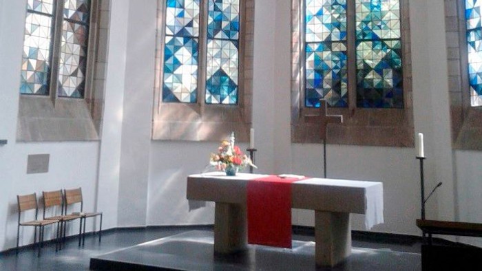 Ein ruhiger Kircheninnenraum mit hohen Buntglasfenstern und einem schlichten Altar, geschmückt mit Blumen und Kerzen.