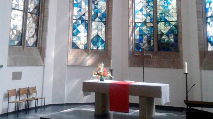 Ein ruhiger Kircheninnenraum mit hohen Buntglasfenstern und einem schlichten Altar, geschmückt mit Blumen und Kerzen.