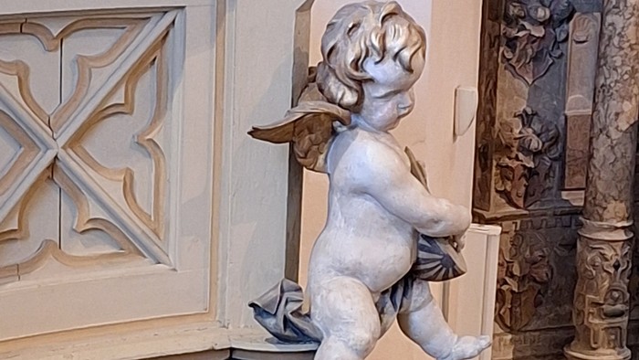 Eine puttenhafte Statue, die auf einem verzierten Sockel mit dekorativen Schnitzereien und architektonischen Details thront.