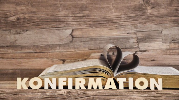 Ein offenes Buch mit herzförmigen Seiten und dem Wort „KONFIRMATION“ darunter auf einem Holzuntergrund.