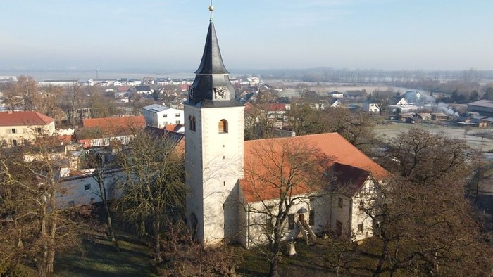 Luftaufnahme einer historischen Kirche mit hohem Turm, die über ein malerisches, weitläufiges Dorf blickt.