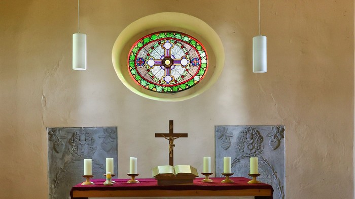 Minimalistischer Kapellenaltar mit Kreuz, Kerzen und einem dekorativen Buntglasfenster sowie einer Sternendeckenverzierung.