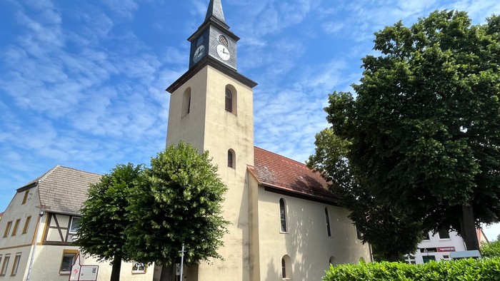 Kirchturm mit Uhr und Kreuz, umgeben von Bäumen und Büschen