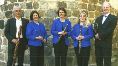 Ein Quartett von Musikerinnen und Musikern in formeller Kleidung, die verschiedene Holzblasinstrumente vor einer Steinmauer halten.