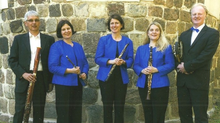 Ein Quartett von Musikerinnen und Musikern in formeller Kleidung, die verschiedene Holzblasinstrumente vor einer Steinmauer halten.