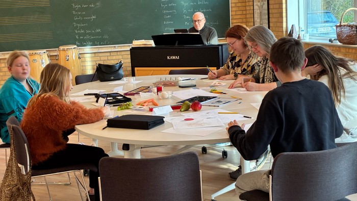 En gruppe samarbejder omkring et rundt bord i et klasselokale, hvor de arbejder med papirer og diskuterer.