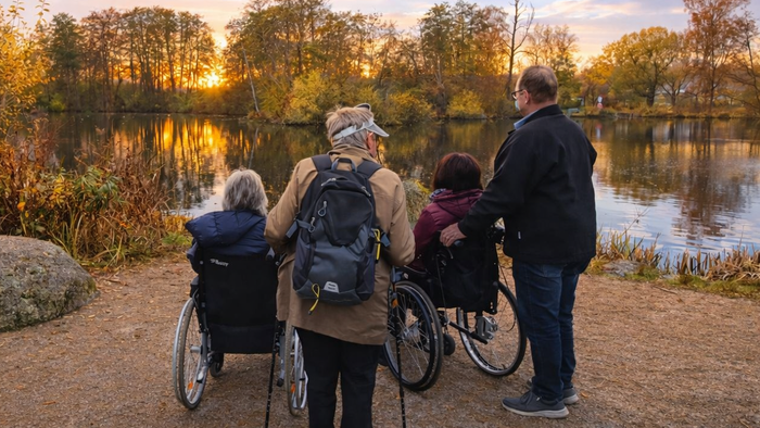 Eine Gruppe genießt einen malerischen Sonnenuntergang am See, wobei zwei Personen im Rollstuhl sitzen und andere Unterstützung leisten.
