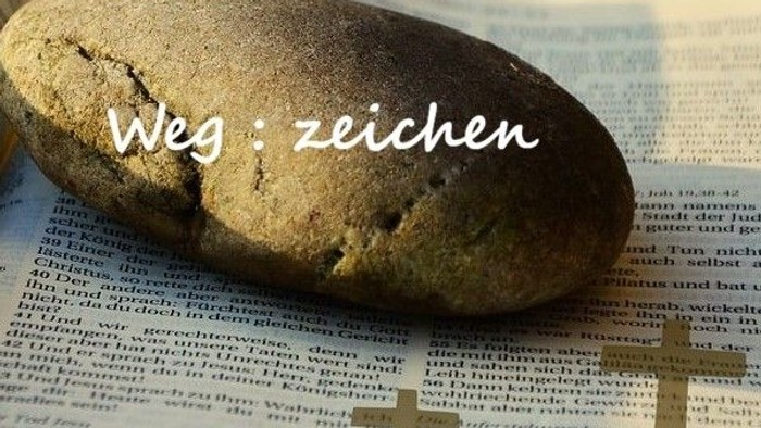 Ein glatter Stein liegt auf einem offenen Buch mit deutschem Text und dem überlagerten Satz „Weg: zeichen“.