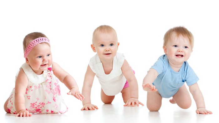 Tre spænde småbørn på hender og knæ, en med rosa hodebånd og blomsterkleid, en i hvid body og en i blå body.