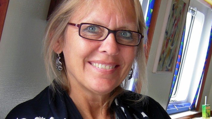 En glad kvinde med briller og en sort jakke med hvide broderede mønstre smiler varmt.