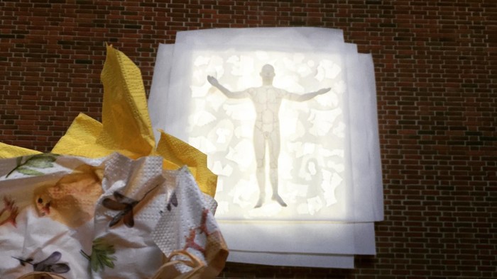 En hånd holder en pakket gave med blomster, foran et lysbillede af en figur og en lille bord med blomster og lys.