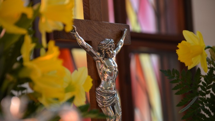 En sølv crucifix med Jesus Kristus på et trækors, omgivet af gule blomster og et farvet vinduesglas i baggrunden