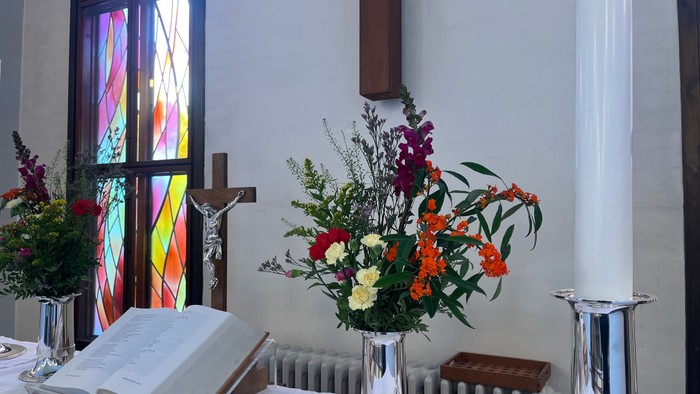 Bord med blomster, bog og lysestage foran farverige vinduesglasning