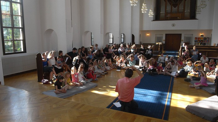En gruppe børn og voksne sidder på yoga-matter i en stor hall med høje vinduer og orgel.