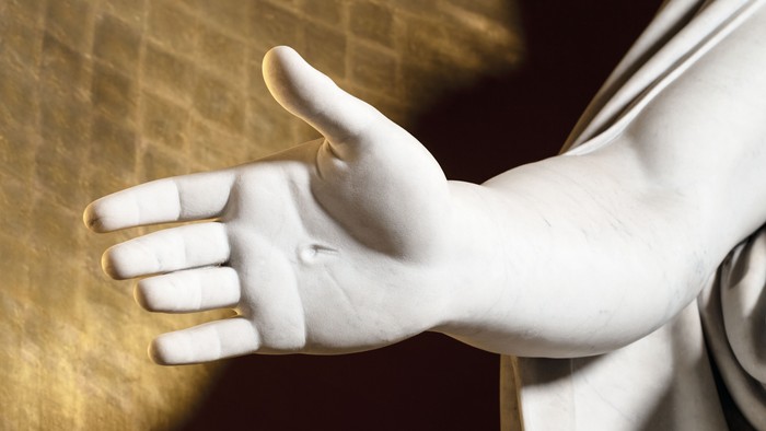 En marmorskulptur af en hånd udstrakt mod en gul mur.