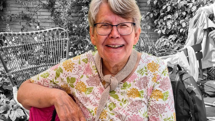 Ældre smilende kvinde i blomstret skjorte, der sidder udendørs nær indkøbsvogne og en barnevogn.