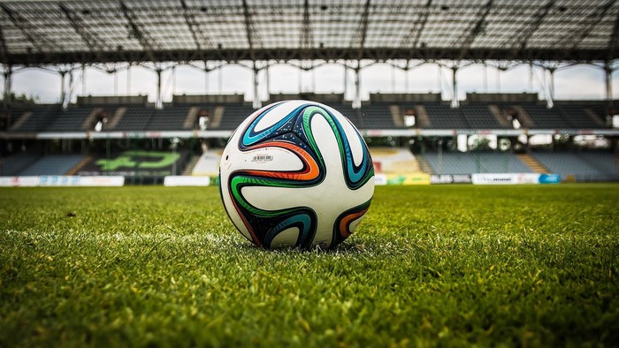 Ein bunter Fußball liegt auf einem saftig grünen Rasenfeld mit Stadionrängen im Hintergrund.