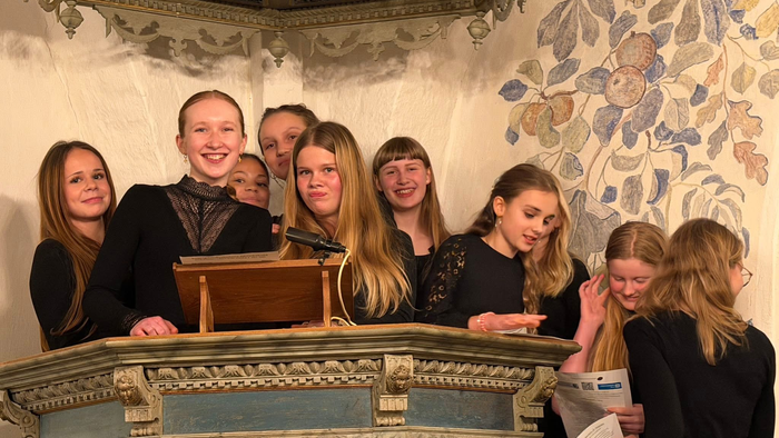 En gruppe unge mennesker samles omkring en prædikestol i et prydeligt rum og ser ud til at forberede sig på en optræden eller tale.