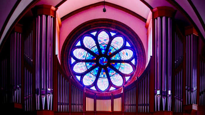 Elegante Pfeifenorgel, gerahmt von einem lebendigen, beleuchteten kreisförmigen Buntglasfenster in einem prächtigen Innenraum.
