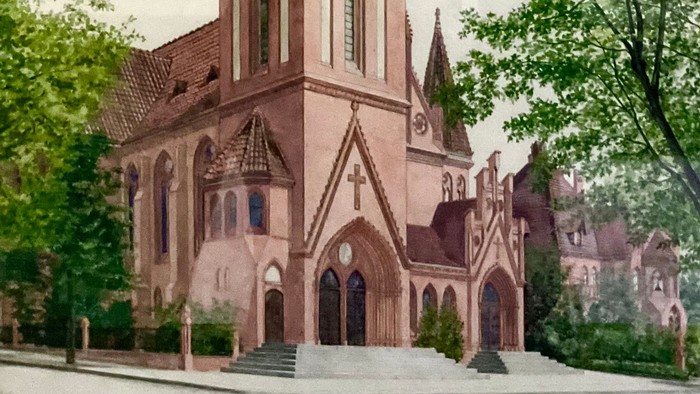 Gotische Kirche mit hohem Turm, eingerahmt von üppigen grünen Bäumen unter bewölktem Himmel.