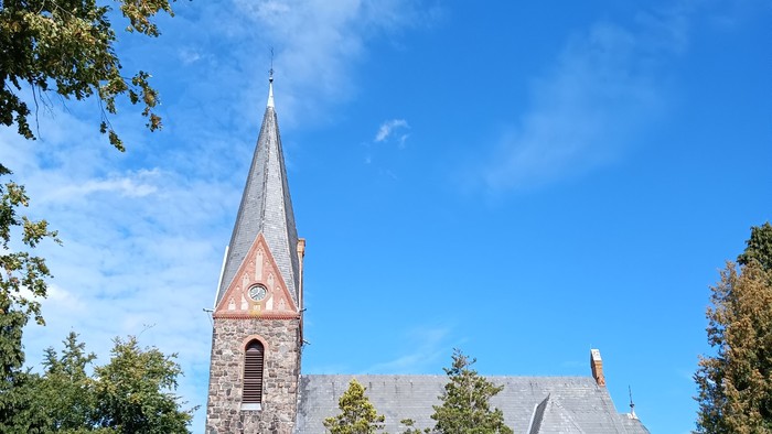 Kirchengebäude mit hohem Turm und spitzem Dach, umgeben von Bäumen und Büschen
