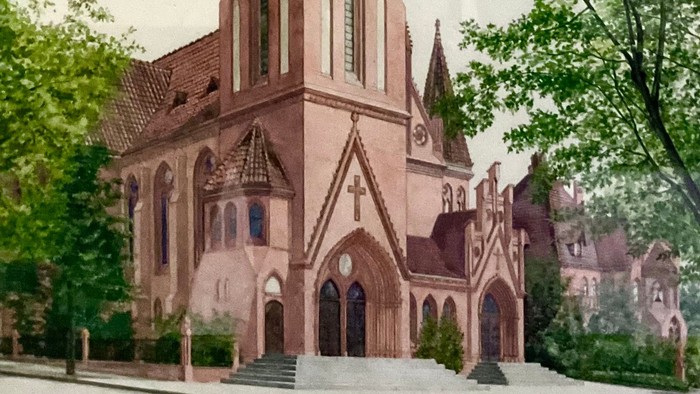 Gotische Kirche mit hohem Turm, eingerahmt von üppigen grünen Bäumen unter bewölktem Himmel.