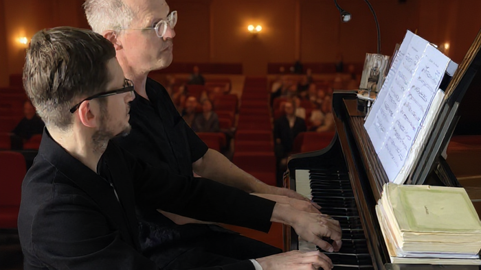 Zwei Musiker spielen gemeinsam Klavier in einem Konzertsaal und konzentrieren sich auf ihre Notenaufführung.