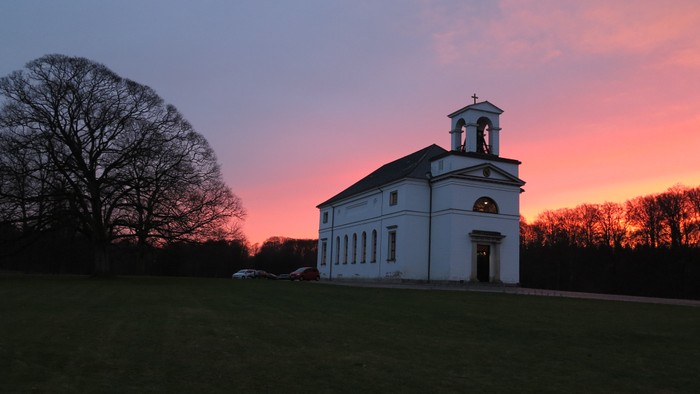 En hvid kirke med tårn og klokke ved solnedgang i en grøn og træfattig landskab