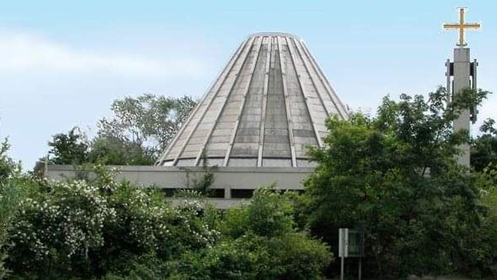 Das Bild zeigt eine moderne Kirche mit einem pyramidenförmigen Dach, umgeben von Bäumen und einem Kreuz.