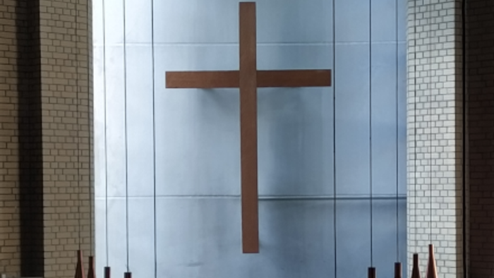 Moderner Kapellenaltar mit einem großen metallischen Kreuz, an dessen Sockel Kerzenhalter angebracht sind.