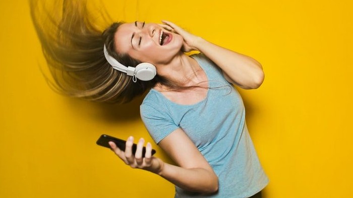 Eine Person, die fröhlich mit Kopfhörern singt und ein Handy hält, vor einem leuchtend gelben Hintergrund.