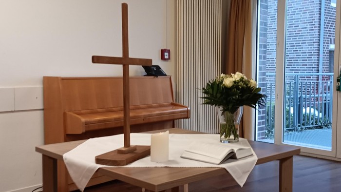 Ein ruhiger Kapellenaltar mit einem hölzernen Kreuz, einer Kerze, einem offenen Buch und einer Blumengesteck neben einem Fenster.