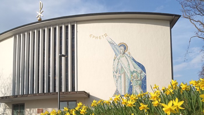 Moderne Kirche mit Wandgemälde eines Heiligen, umgeben von leuchtend gelben Osterglocken im Frühling.
