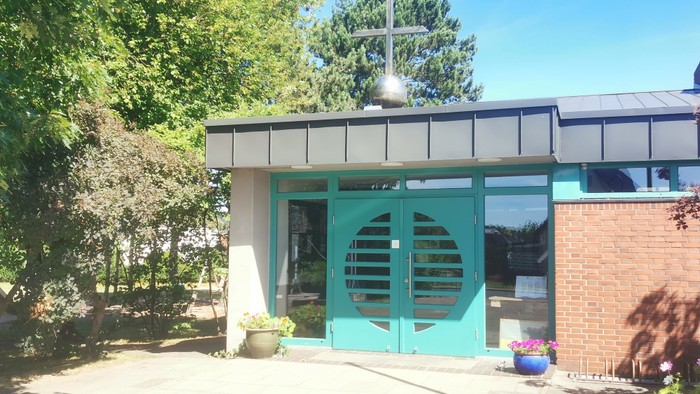 Kleine Kirche mit grünem Vordach und Kreuz auf dem Dach, umgeben von Bäumen und Blumen.