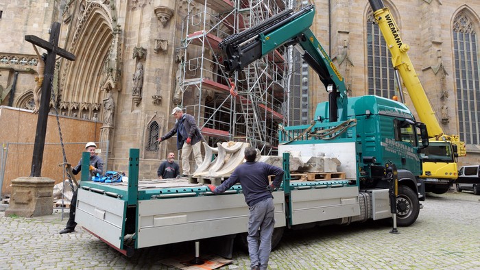 Ein Baustellenwagen mit Kranaufbau steht vor einer Kirche und hebt Bauteile an.