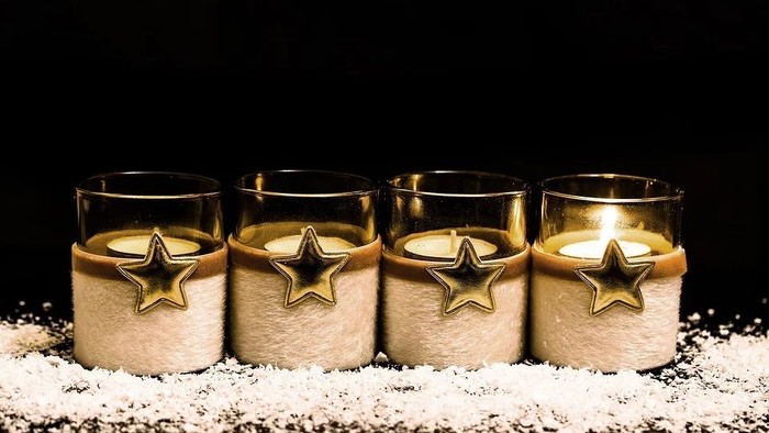 Vier brennende Kerzen in Glashaltern mit Sternenmotiven, aufgestellt auf einer schneebedeckten Fläche.