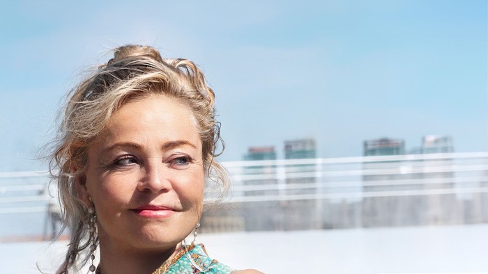 Kvinde med krøllet hår sat op, smilende udendørs nær byens skyline.